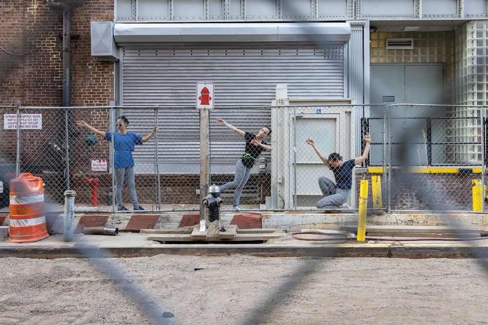 Three dancers in a row create a tableau while grasping onto a metal link gate 